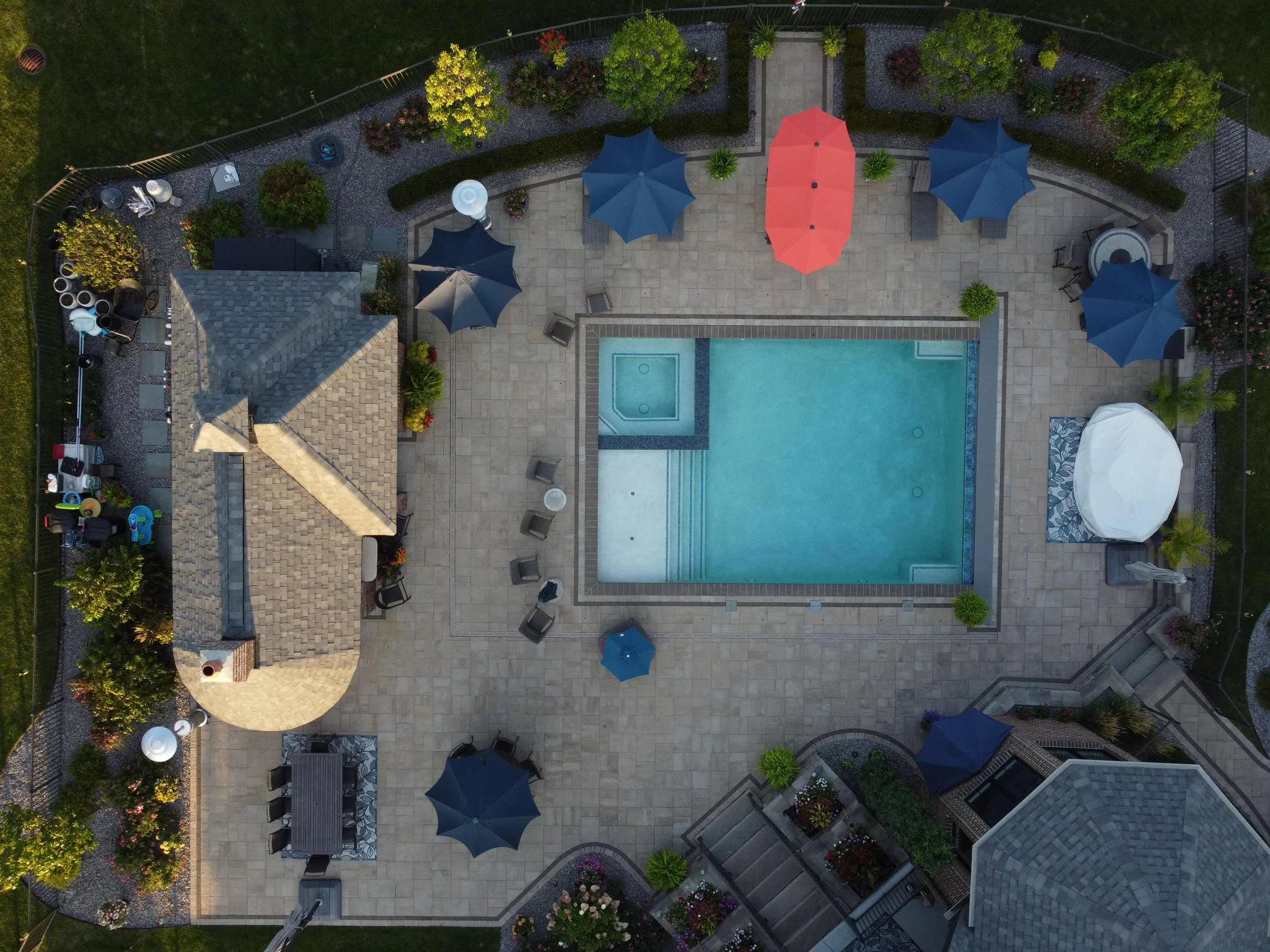An aerial view of a backyard swimming pool area with blue and red umbrellas, outdoor furniture, a hot tub, a gazebo, landscaping, and a surrounding stone patio.