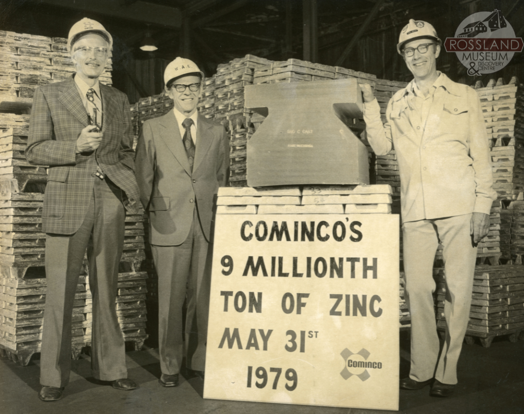 Black and white photograph of three men standing for a promotional photo with large zinc bar sitting on a large pile of zinc bricks. More bricks are in the background in large stacks