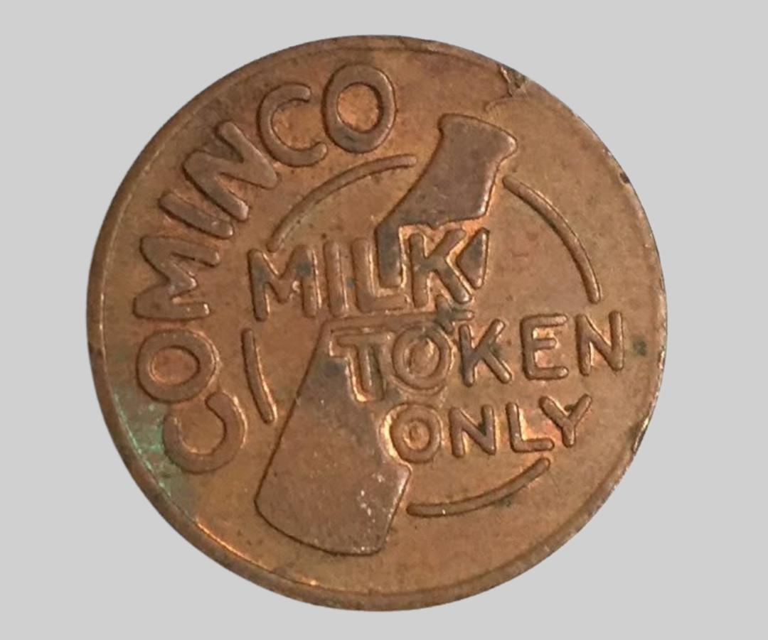 Copper-coloured coin on a grey background with "Comino Milk Token Only" and a milk bottle on the front of coin