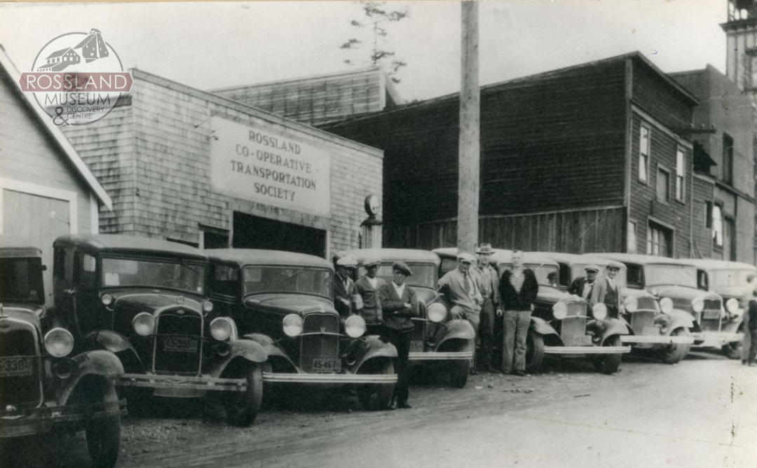Black and white image of eight 1930s era cars in front of several buildings