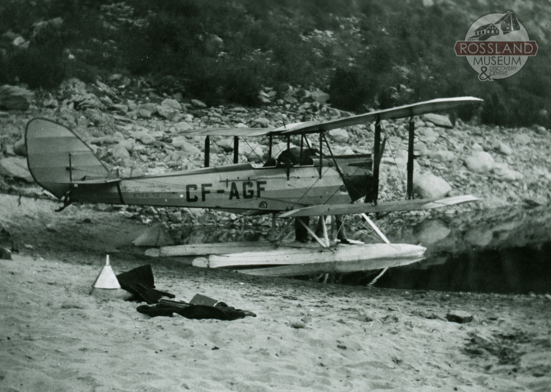 Black and white photograph of a 1930s era float plane on a waterbody by the beach with a rocky hillside in the background