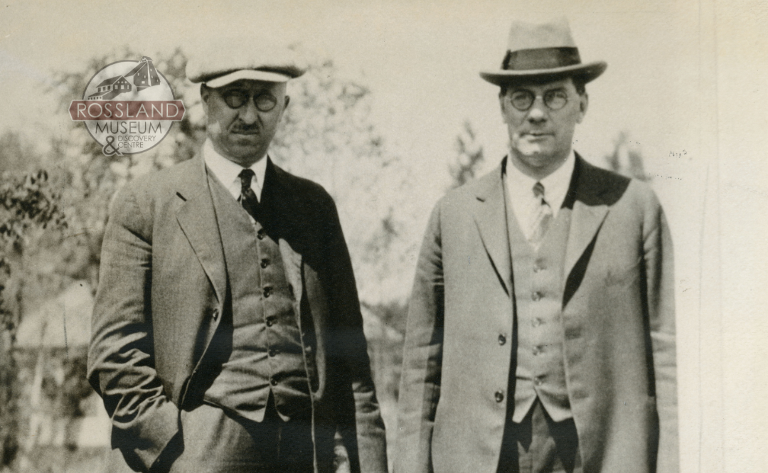 Black and white photograph of two men in three-piece suits with trees in the background