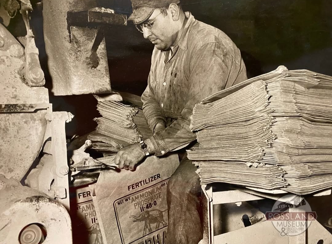 Black and white photograph of man surrounded by piles of paper fertilizer bags