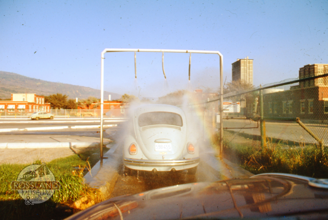 Colour photograph of a blue VW bug going through single frame car wash with industrial buildings in the background