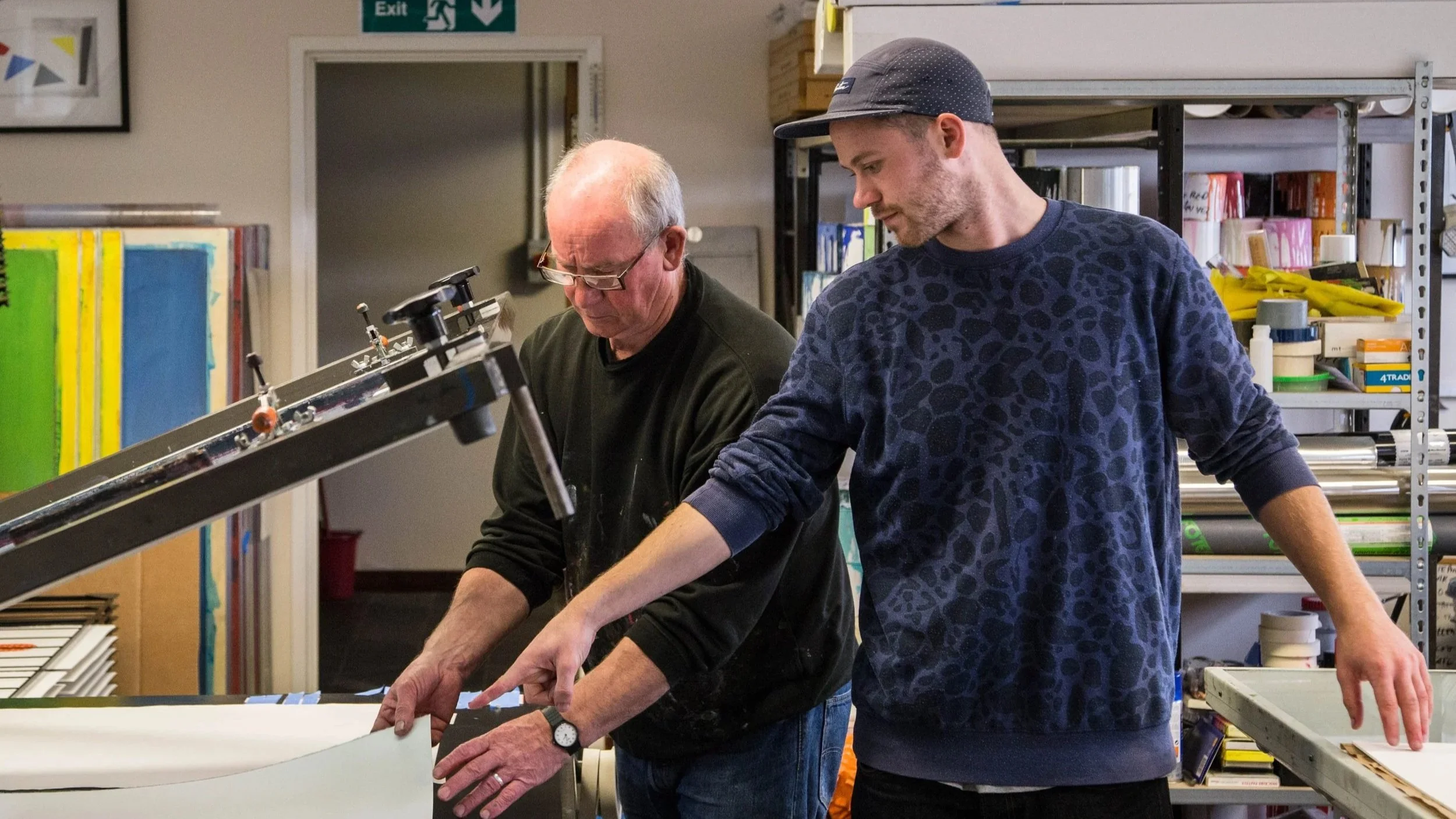 Two men working together in a print studio workshop, examining materials on a table, with shelves of supplies in the background. It features Kip Gresham and his son Tim Gresham