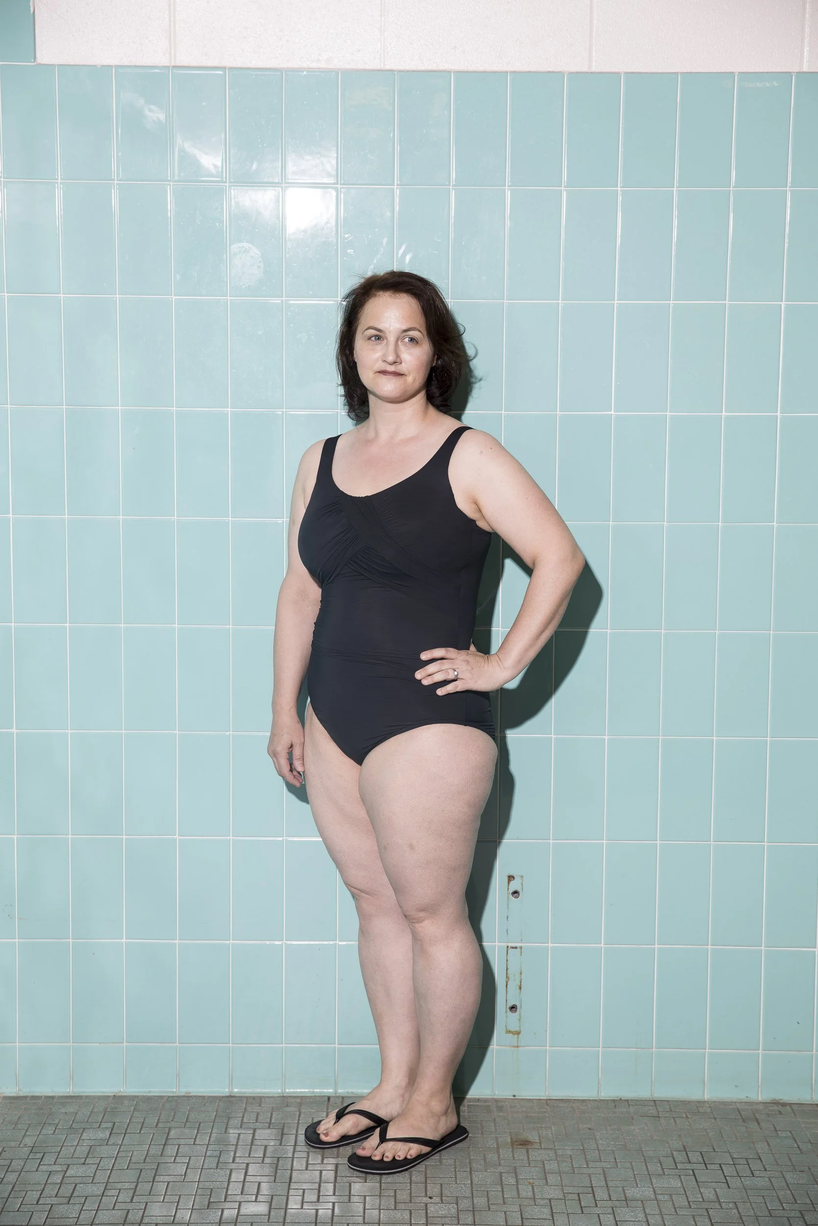 A woman standing in front of a tiled wall, wearing a black swimsuit and flip-flops, with her hand on her hip.