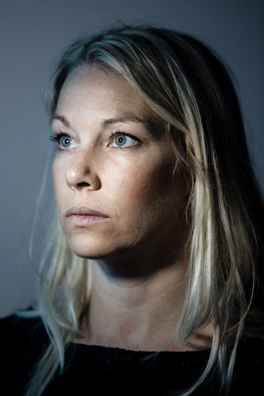 A woman with blonde hair looking off to the side, with bright blue eyes, wearing a black top, against a dark background.