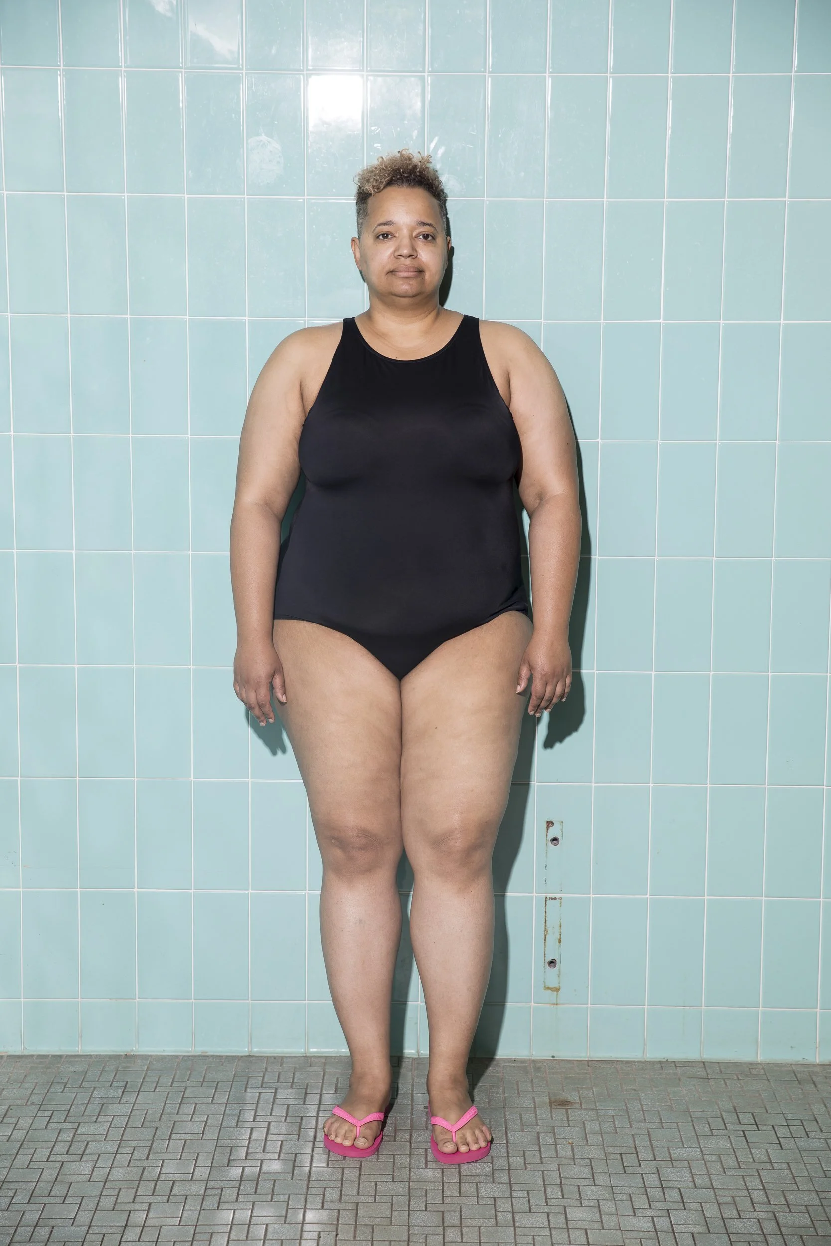 A woman in a black swimsuit stands barefoot on a tiled floor against a teal tiled wall, looking at the camera with a neutral expression.