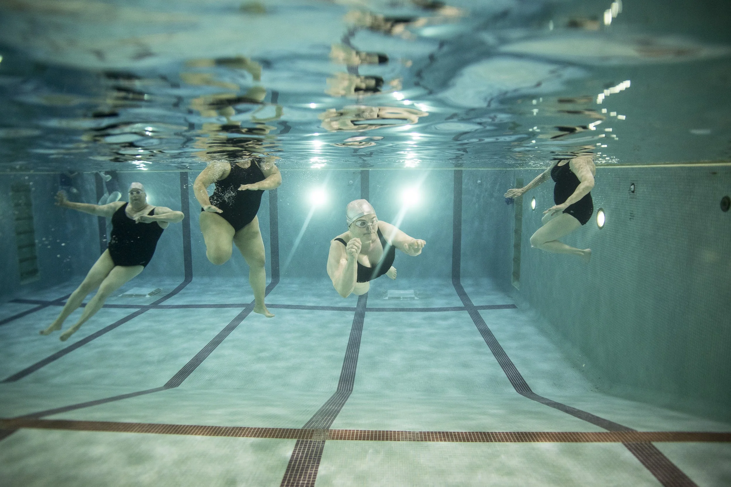 Four women in swimsuits swimming underwater in a pool.