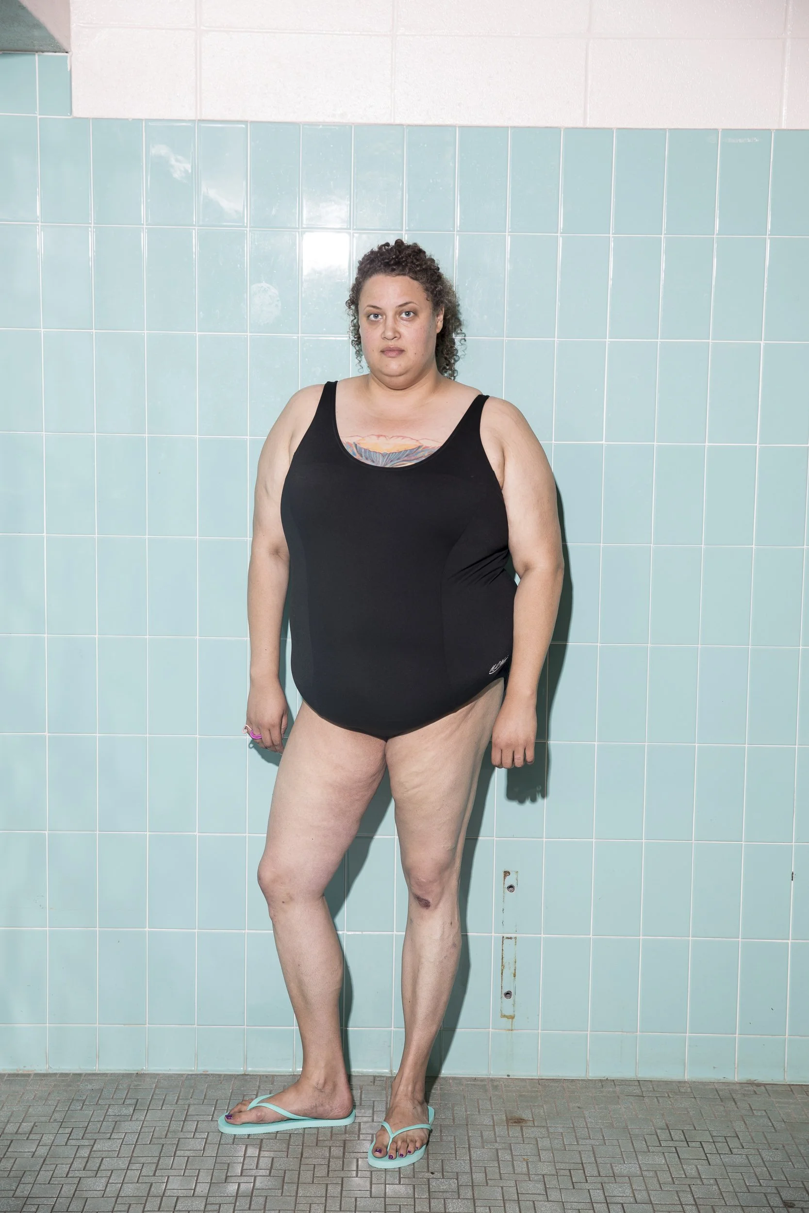 A woman standing in front of a tiled wall, wearing a black swimsuit and flip-flops, with her arms at her sides.