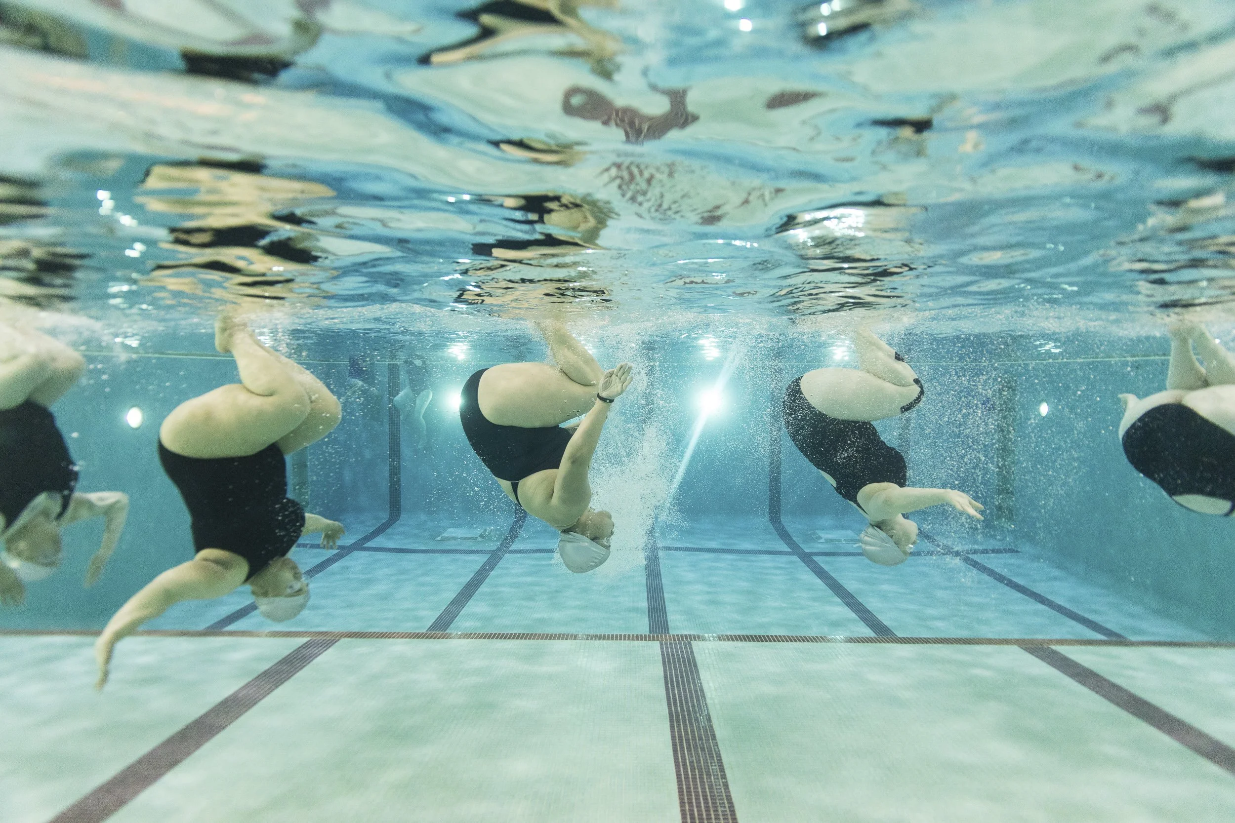 Swimmers practicing synchronized swimming in a swimming pool, submerged underwater with swimmers wearing black swimsuits and white swim caps.