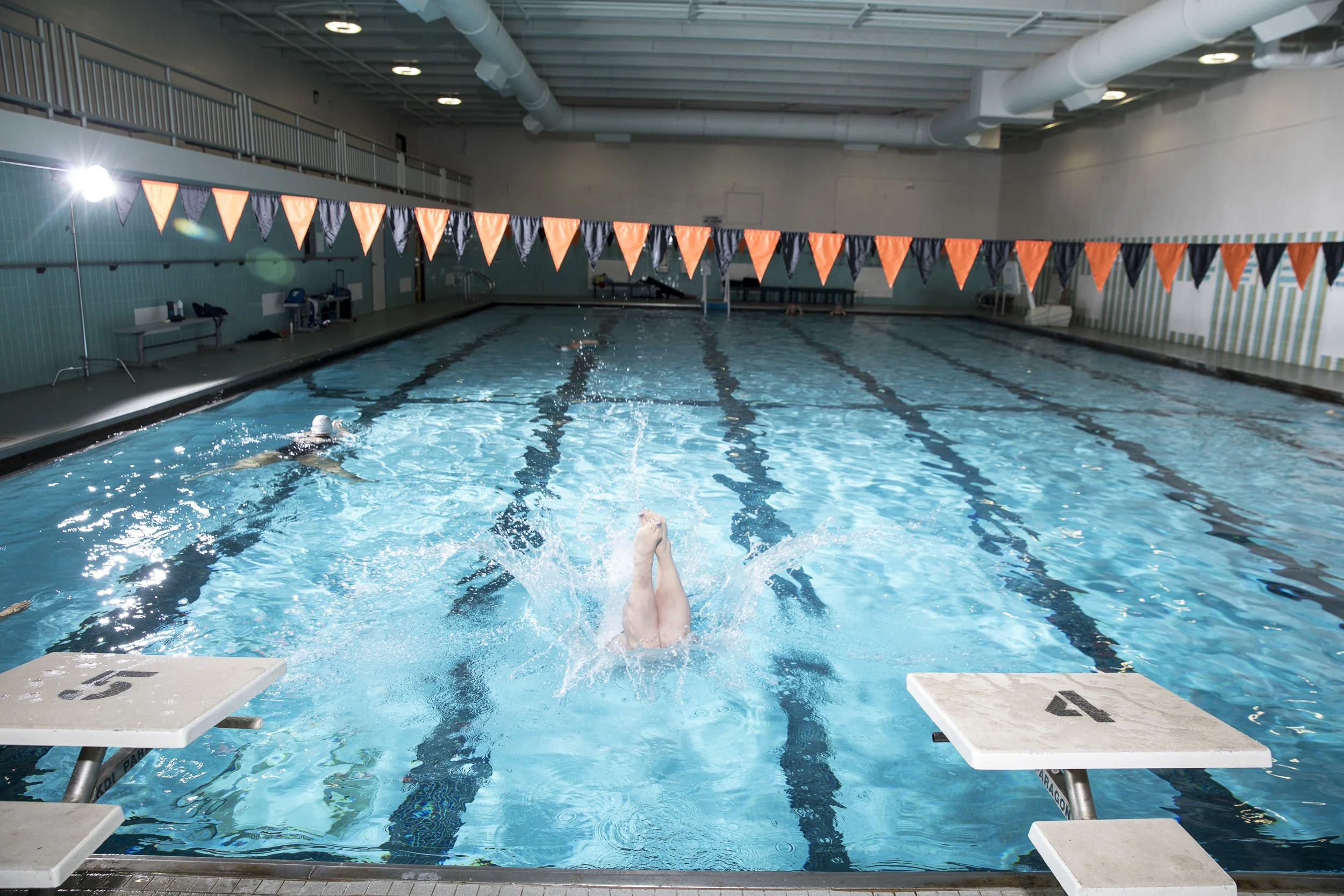 Indoor swimming pool with a swimmer diving or jumping into the water, starting a swim. Pool lanes are marked with black lines, and orange and black triangular flags hang above. Starting blocks numbered 4 and 5 are at the edge of the pool.