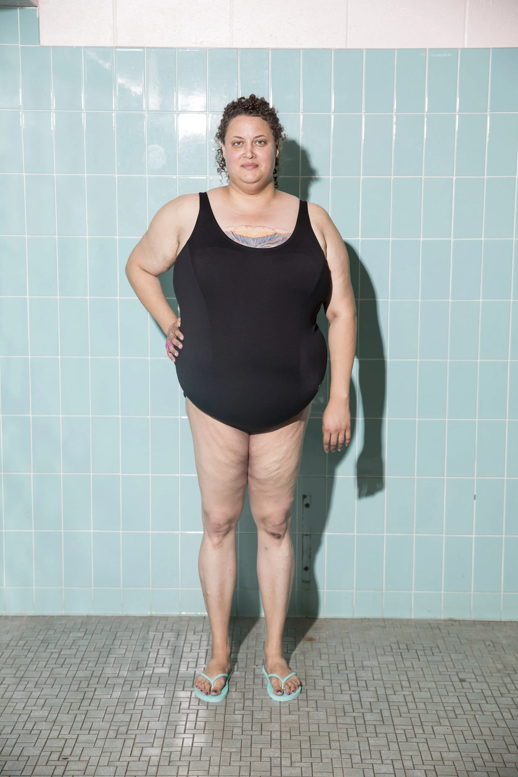 A woman with curly hair wearing a black swimsuit and teal flip-flops standing in front of a light blue tiled wall.
