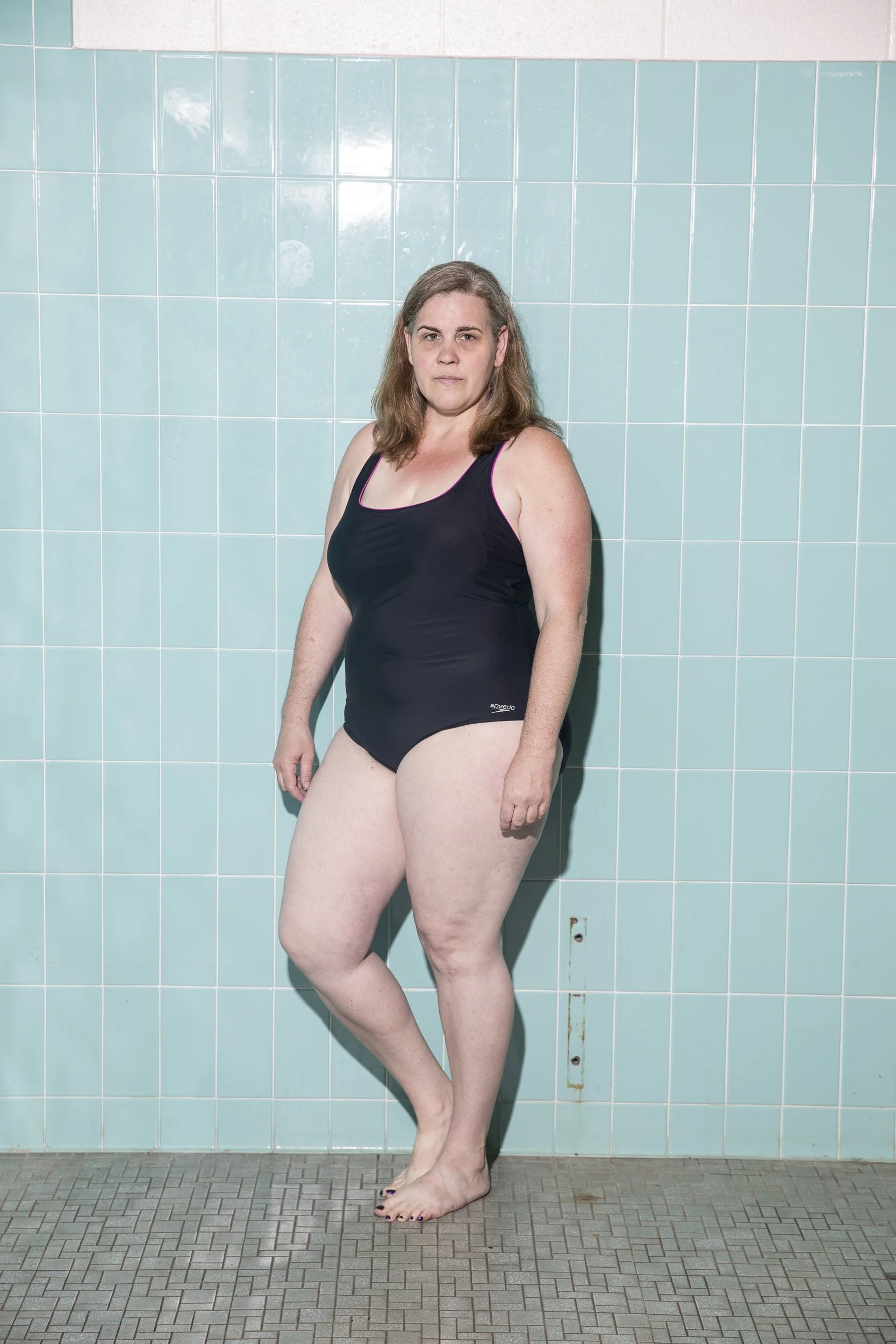 A woman in a black swimsuit stands in a swimming pool area with light blue tiled walls and gray tiled floor.