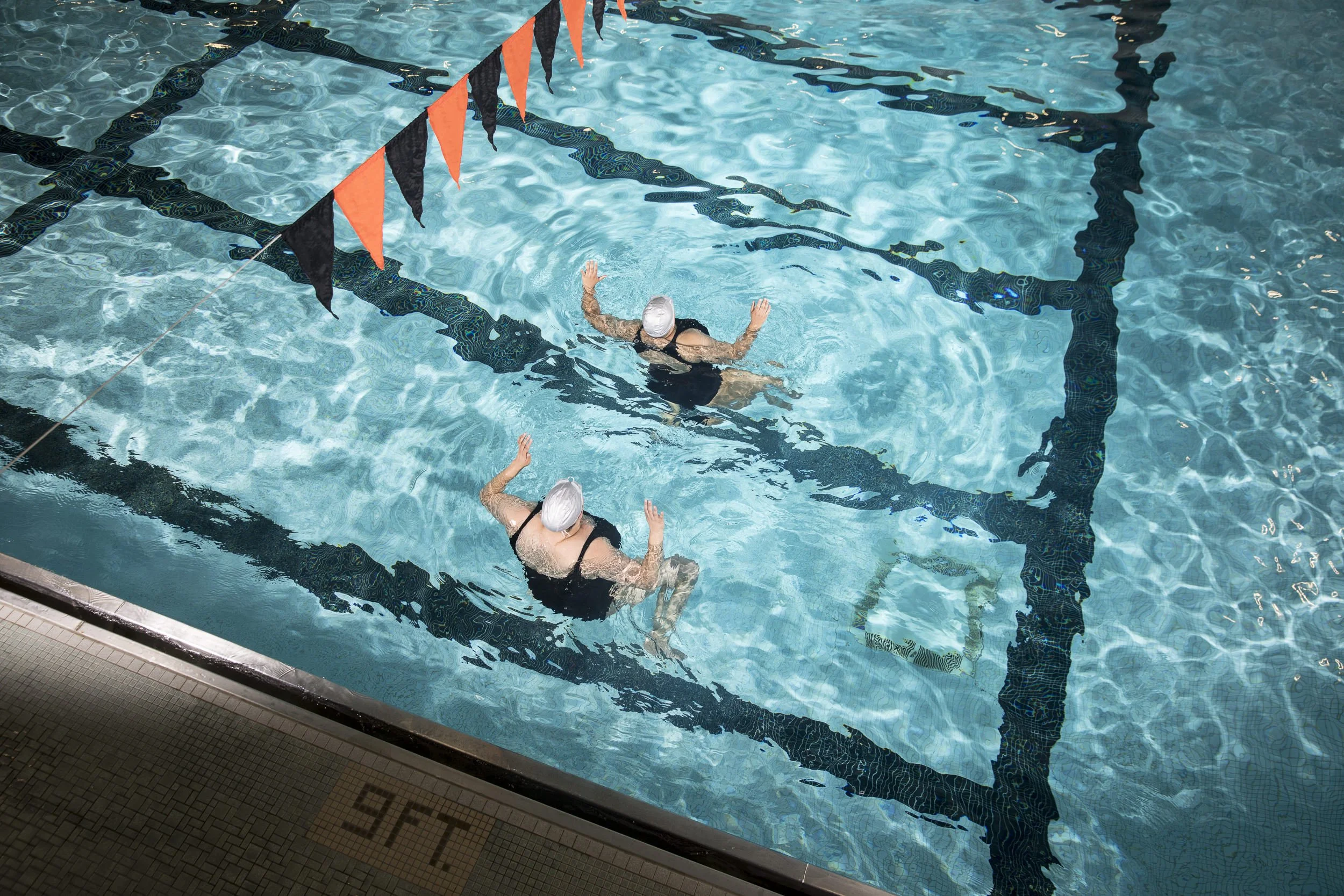 Two swimmers in a swimming pool practicing flip turns during a swim meet, with lane lines and orange and black pennant banners hanging above the pool.