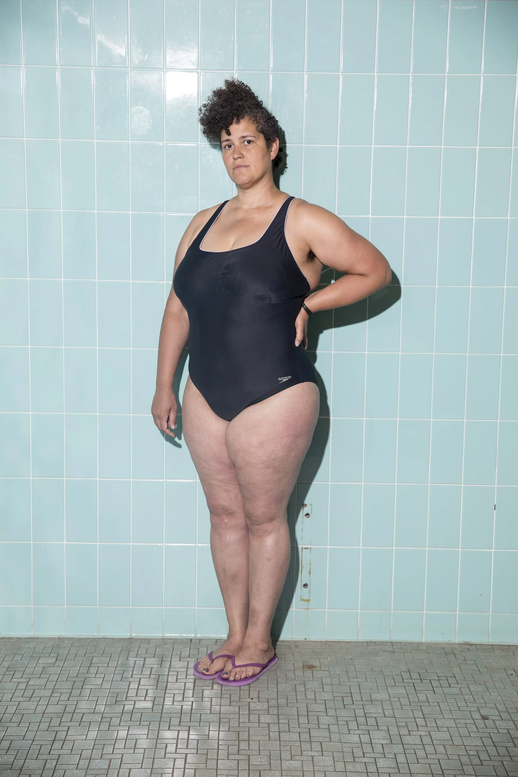 A woman standing in a tiled room, wearing a black swimsuit and purple flip-flops, with one hand on her hip, looking at the camera.