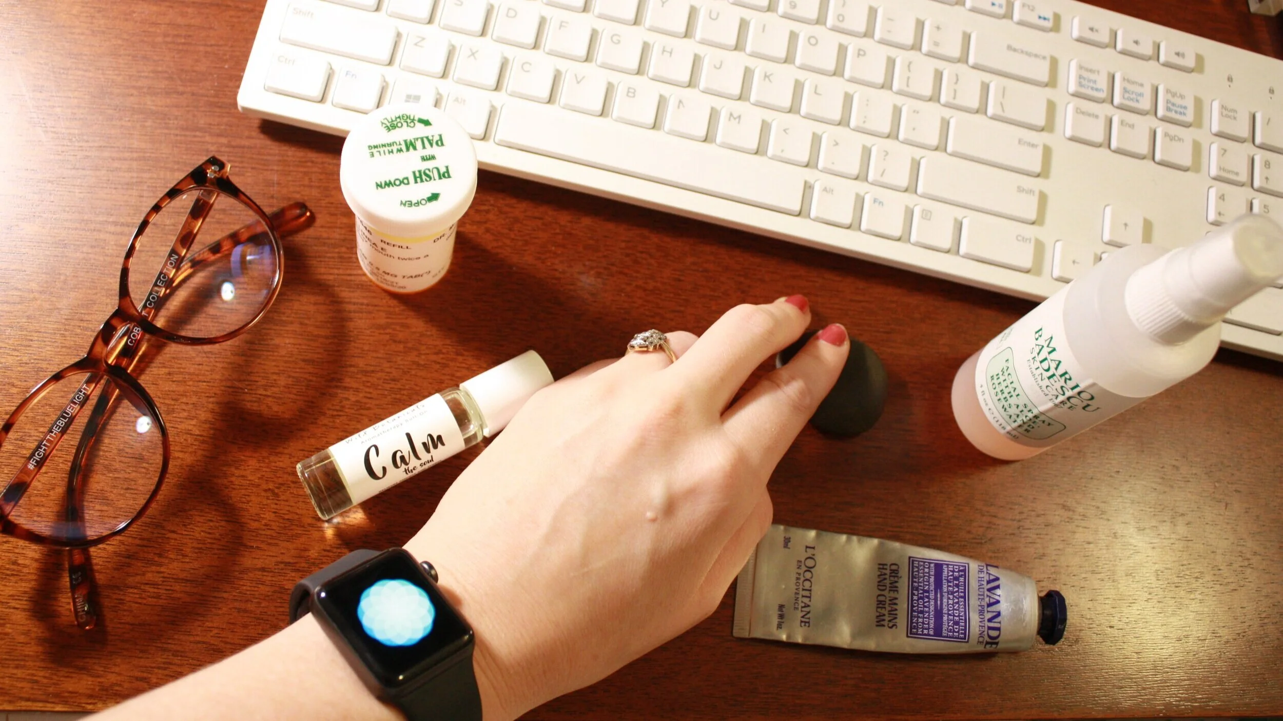 A cluttered wooden desk with glasses, a hand wearing a smartwatch, a perfume bottle labeled 'Calm the soul,' a small container of Push Down medication, a bottle of Mariou Blemish Facial Spray, a tube of hand cream, a black stress ball, a computer keyboard, and other skincare products.