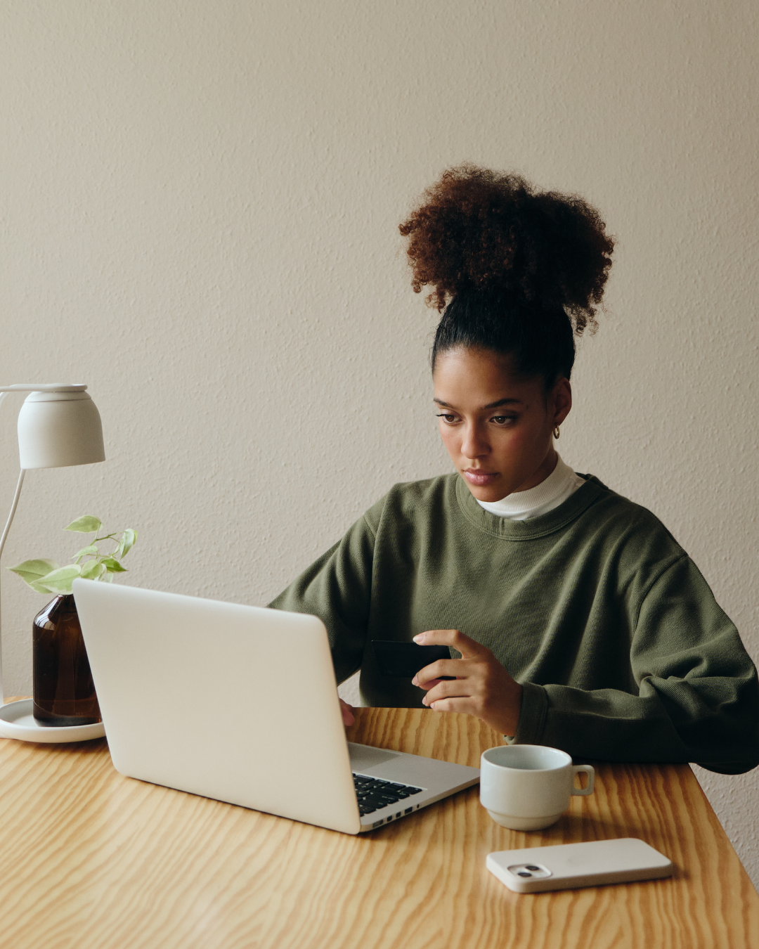 A medium skinned woman with black curly hair and a green sweater sits at a desk with a laptop