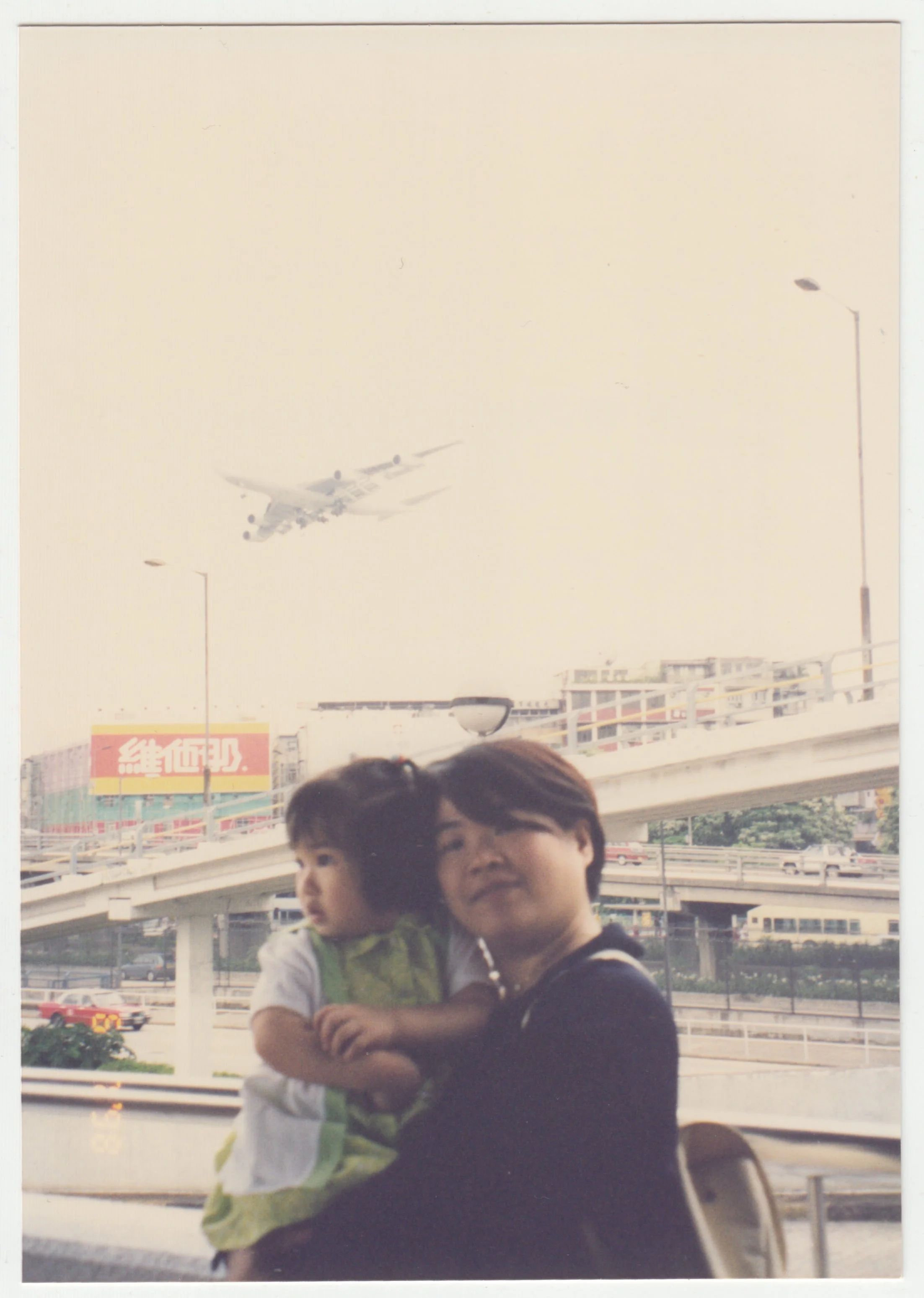 Mum & Charlotte at Kai Tak Airport, Hong Kong, taken on 6 July 1998. There is a plane flying behind us, with the old Vitasoy sign.