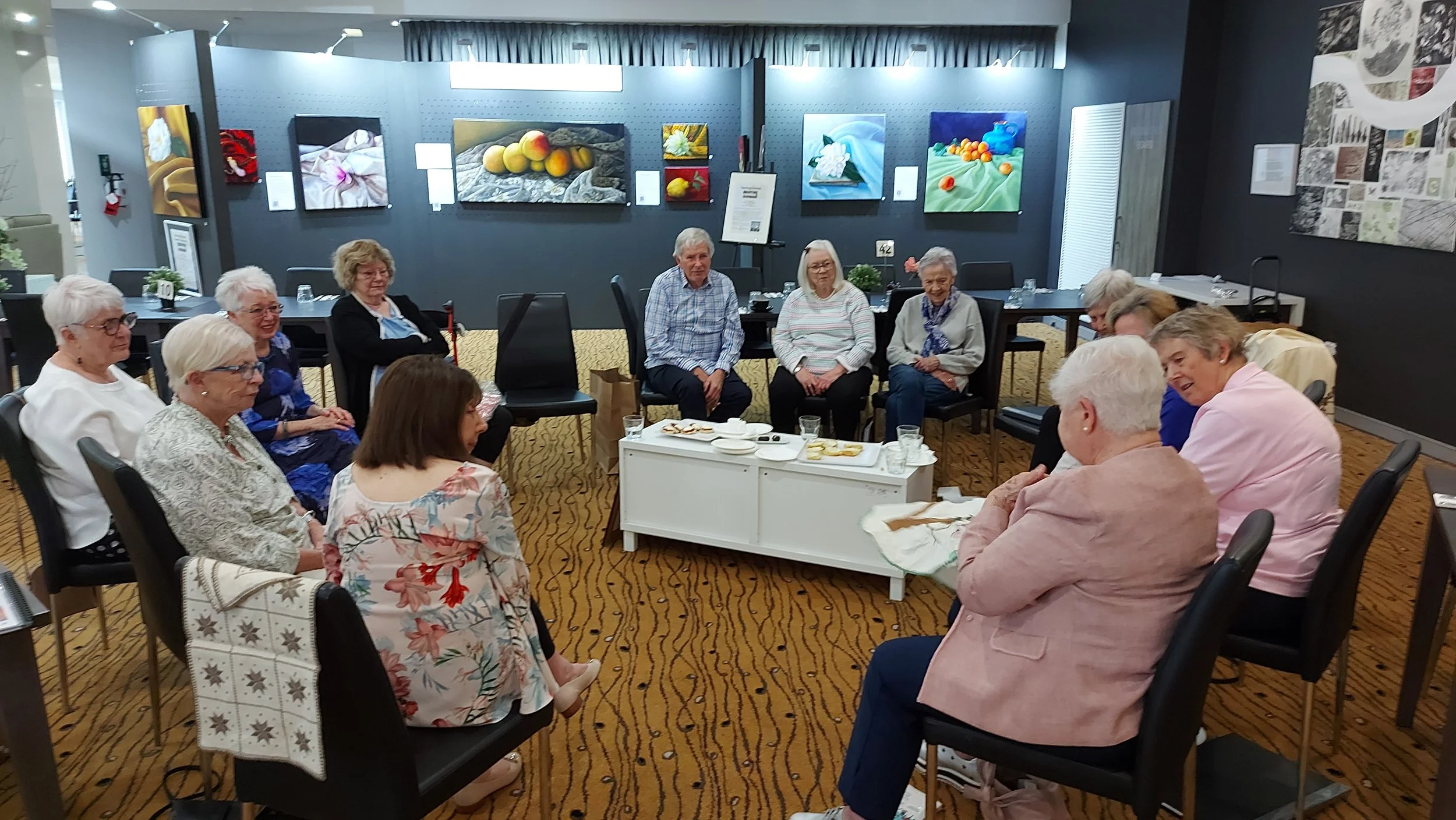 elderly men and women sit in a yarning circle with a painting exhibition in the background
