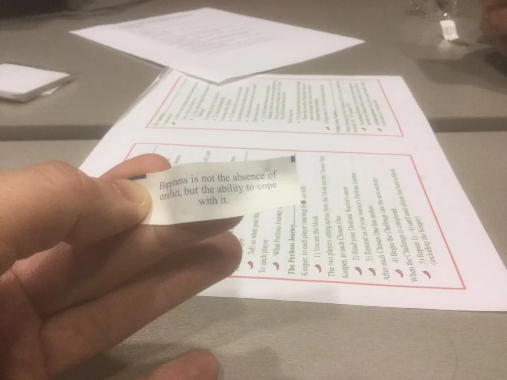Mendez’s hand with a fortune that reads, “Happiness is not the absence of conflict, but the ability to cope with it.” Behind him on the table are some game materials, one of which is a rules cheat with green text and red borders like a Chinese menu. And chili pepper bullet points.