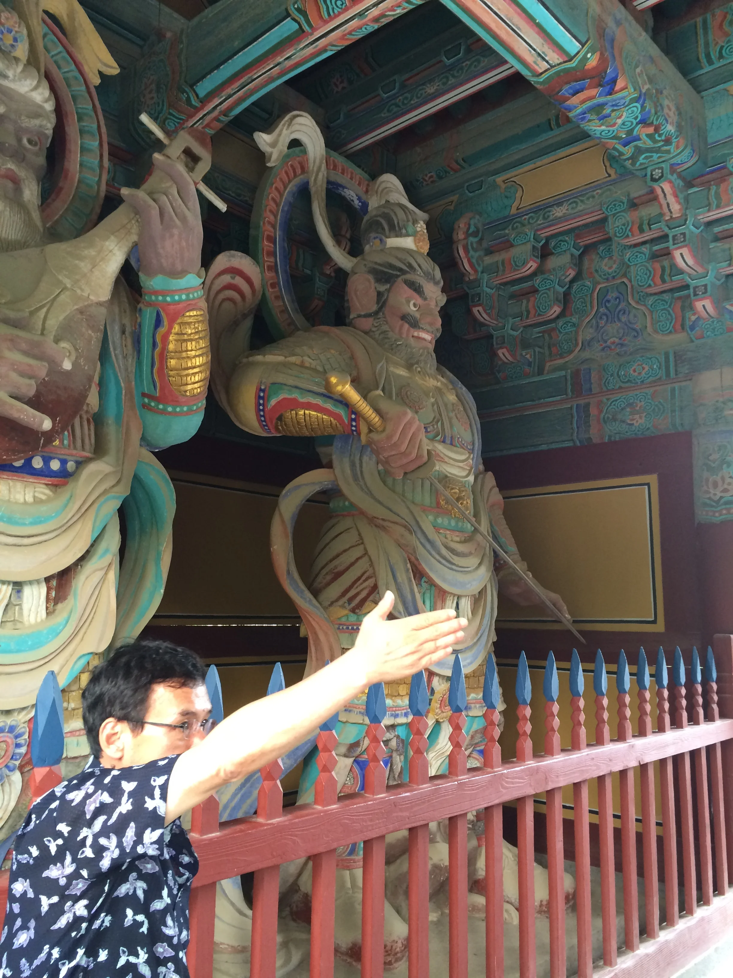 Tour guide Sam Gim stands before towering, colorful statues of guardian gods in a Korean Buddhist temple. Dhṛtarāṣṭra/Jiguk’s on the left playing his lute, Virūḍhaka/Jeungjang brandishing his sword on the right.