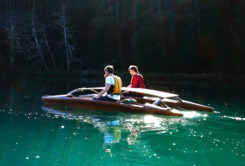 A couple share a two-part canoe on a green river float by forest.