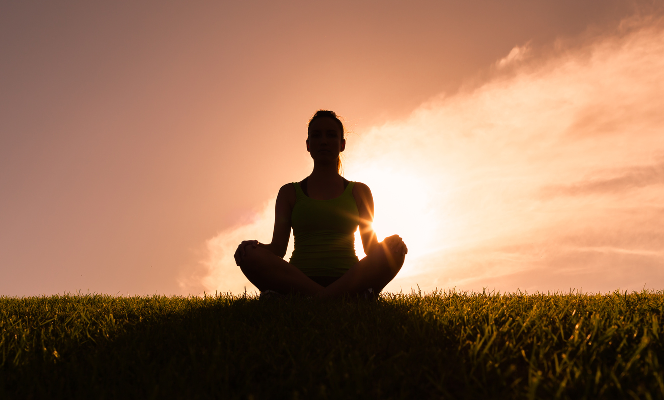 woman sitting in lotus position on a patch of grass, backlit by the sun