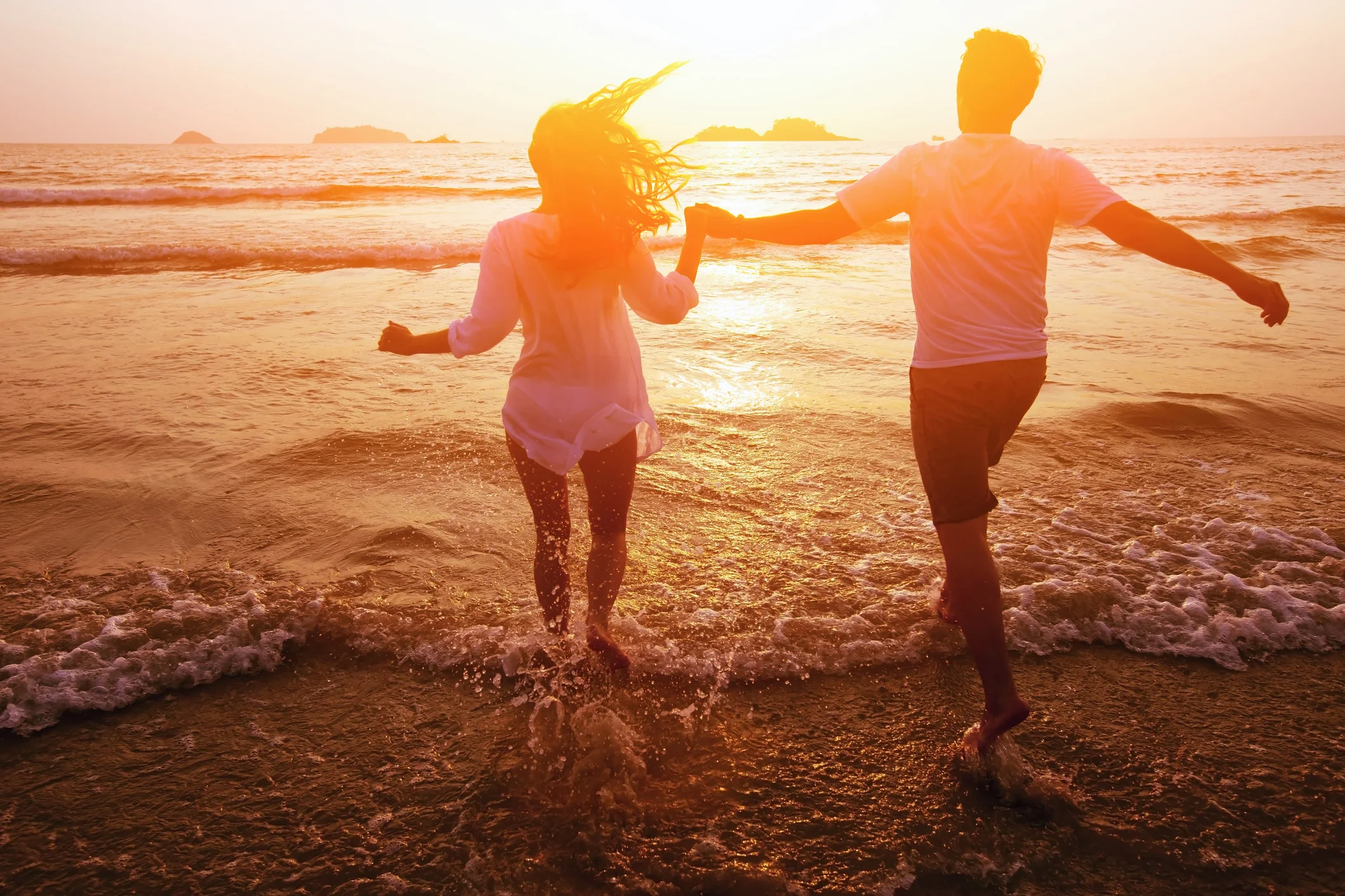 a man and woman hold hands in the late evening sun and walk into the surf