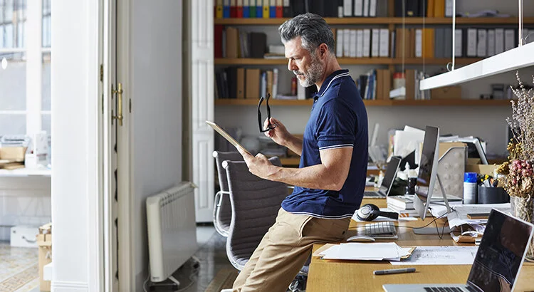 man in an office with bookcases looking at poper while holding reading glasses