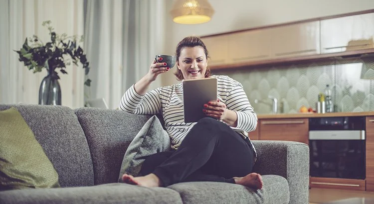 woman looking at iPad on couch while drinking coffee and smiling