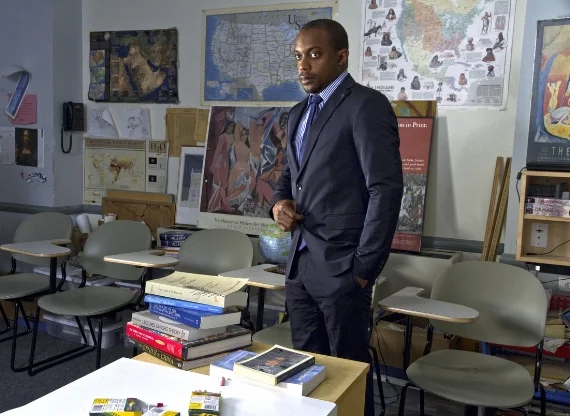 Elia Alba, The Professor (Hank Willis Thomas), 2014. Courtesy of the artist. [image description: a man in a suit is standing facing left at the top of a class room where there are chairs and maps on the wall, he stands behind a desk where there are …