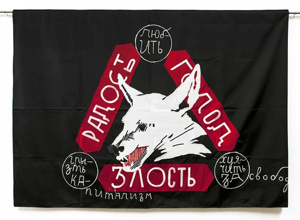 Chto Delat, “Hunger, Anger, Joy,” 2011. Photo: KOW. Courtesy of Chto Delat and KOW, Berlin | Madrid. [image description: white dog with fangs bared is set in a red triangle of text in cyrillic font, with the words in the title, all on a black backgr…