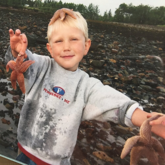 child holding two starfish in his hands and one on his head with a shirt that says Pembroke, ME on it