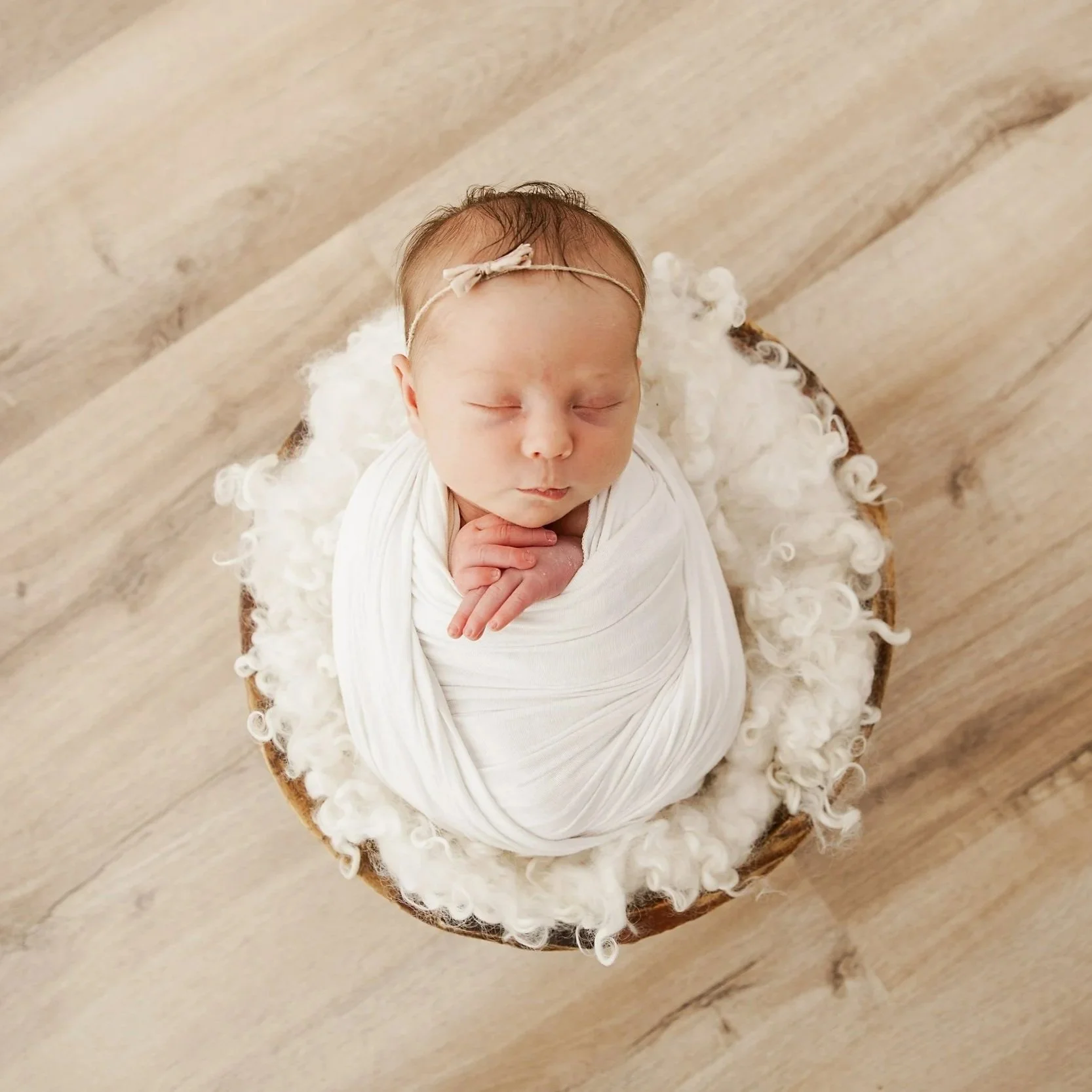 A sleeping newborn baby wrapped in a white blanket, lying in a round basket with fluffy white lining, placed on a wooden floor.