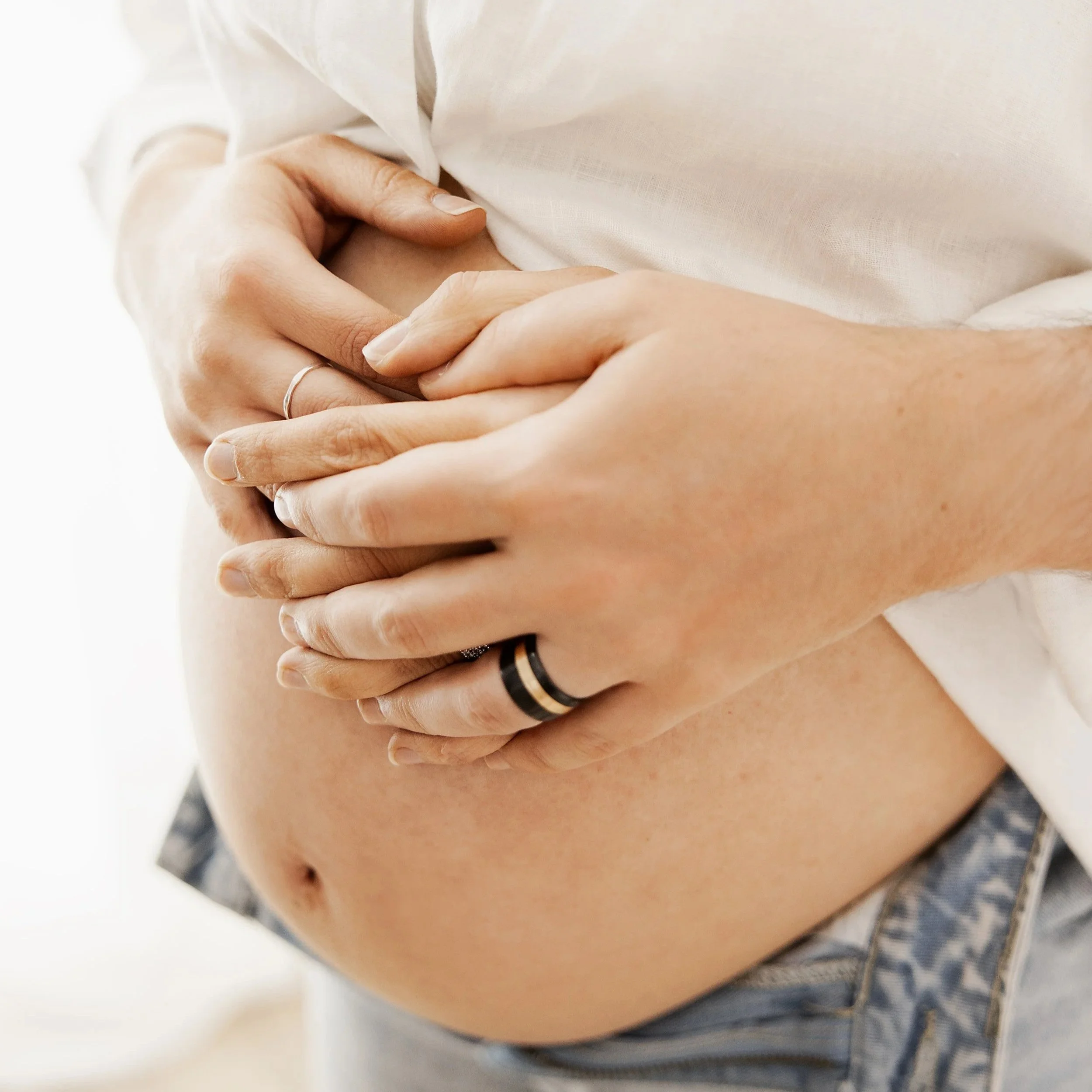 Close-up of a pregnant person's belly with hands gently resting on it, wearing rings on fingers.