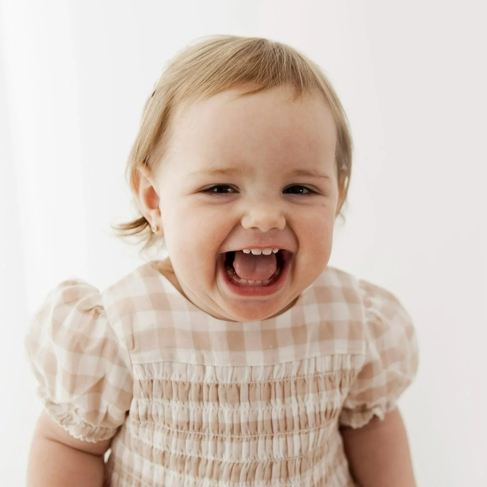 A happy young girl with short blonde hair, wearing a peach-colored checkered dress, laughing with her mouth open, against a plain white background.