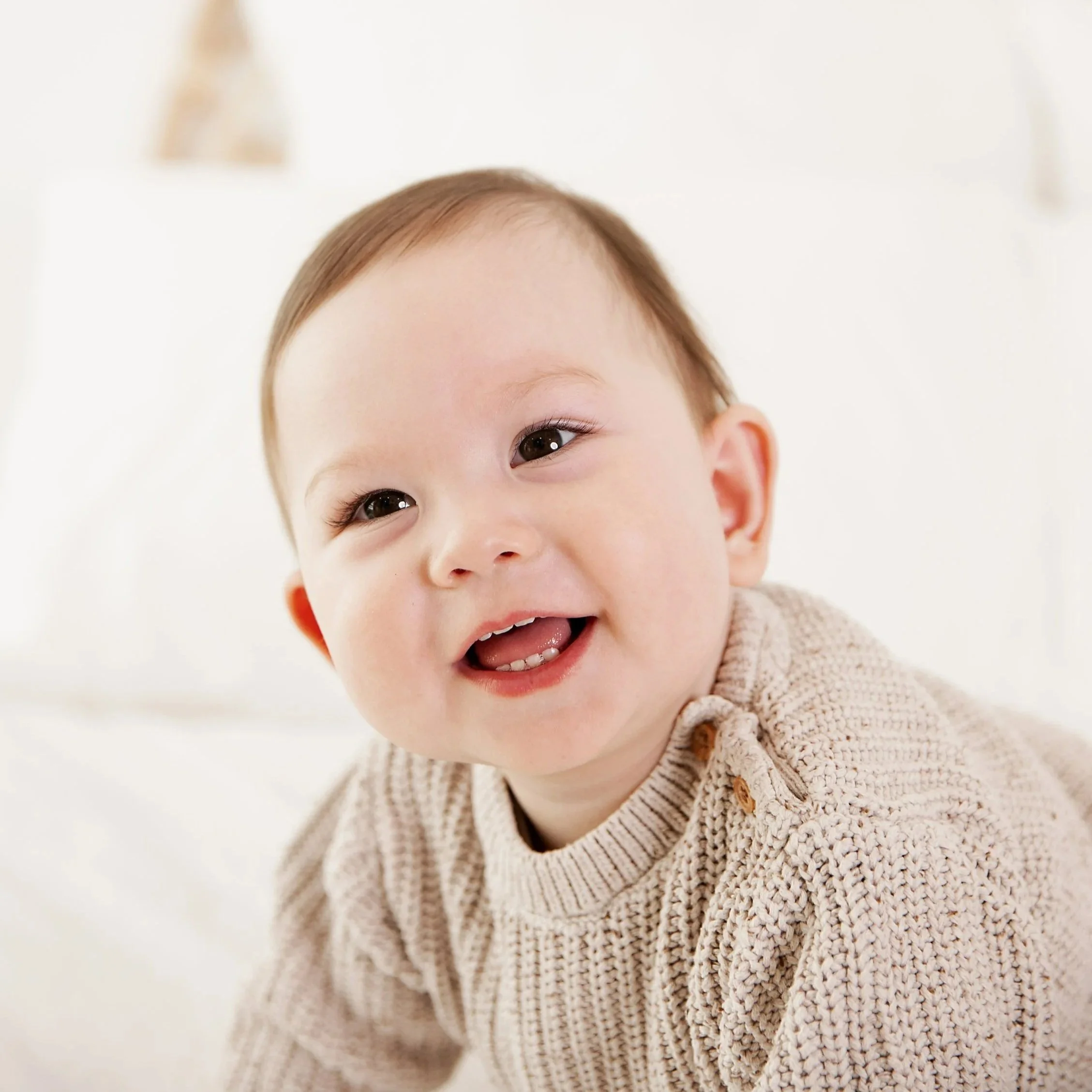 Close-up of a smiling toddler with light brown hair, wearing a beige knitted sweater, in a bright, minimalistic setting.