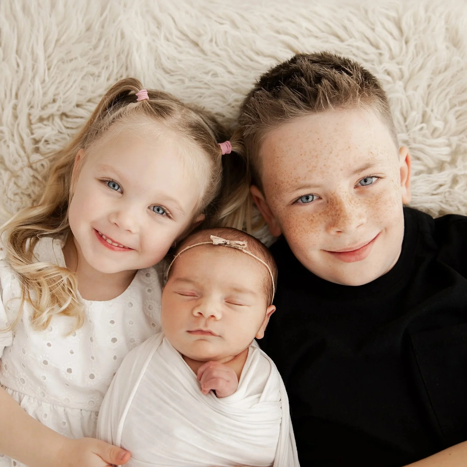 A family of four with a young girl, a teenage boy, a newborn baby girl, all lying on a fluffy white blanket, smiling at the camera.