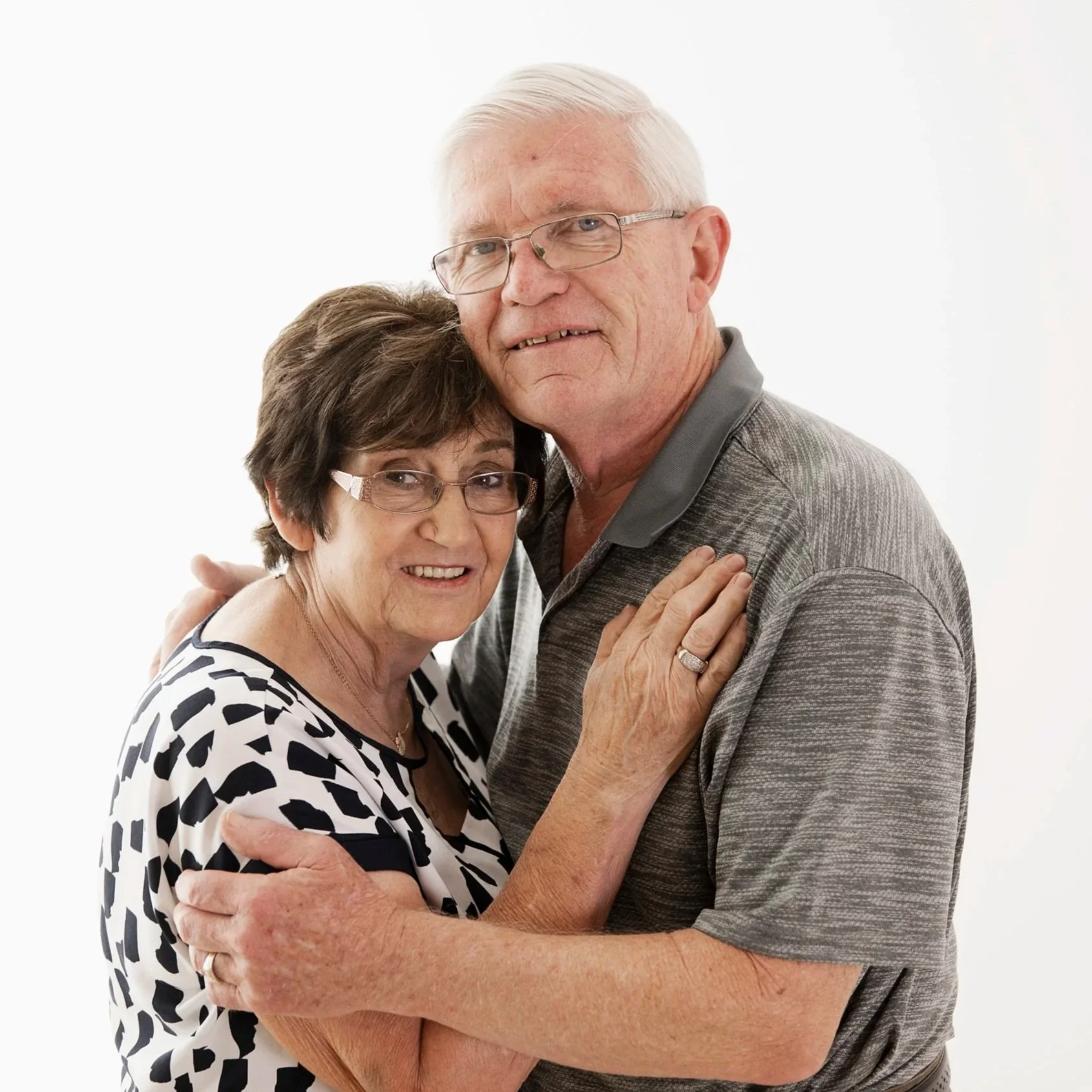 An elderly couple hugging and smiling at the camera against a plain white background.