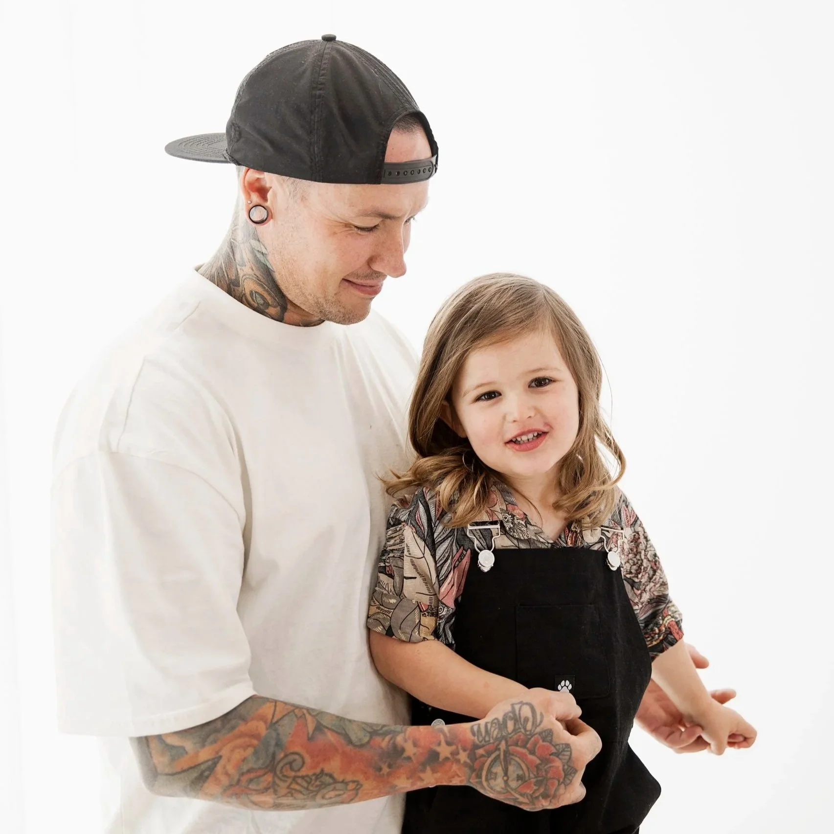 A man with tattoos and piercings wearing a backward black cap and a white t-shirt holding a young girl with long brown hair, a floral shirt, and a black overall, against a plain white background.