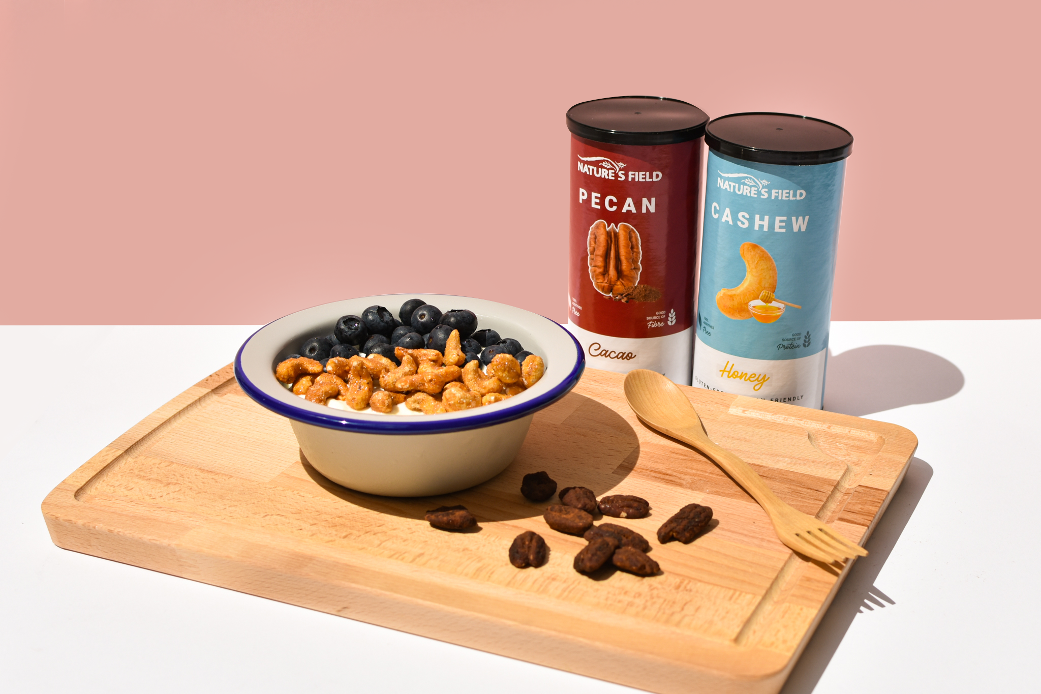 Nature's Field cacao pecans and honey cashews consumed with fresh blueberries in a yogurt bowl with a wooden spoon.