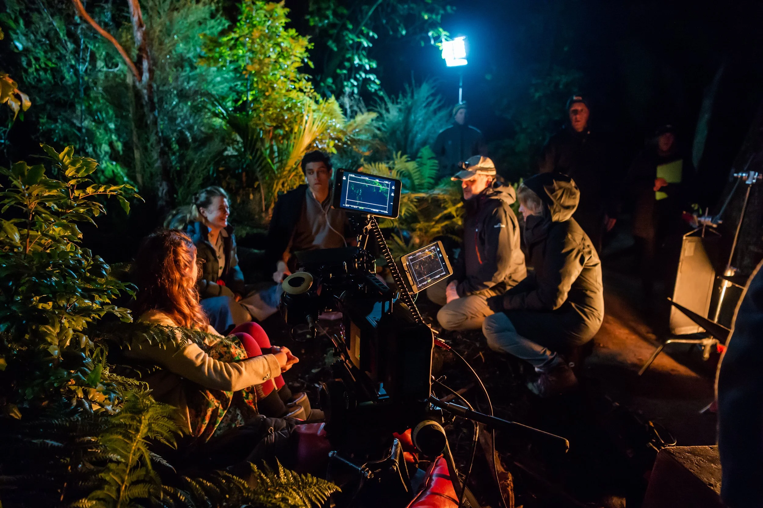 Actors talking with the directors gathered outdoors at night, surrounded by lush greenery, with filming equipment including cameras and monitors set up, indicating a filming session in a forested area.