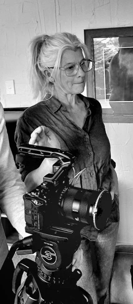 Filmmaker stands next to a camera on a tripod in an indoor setting.