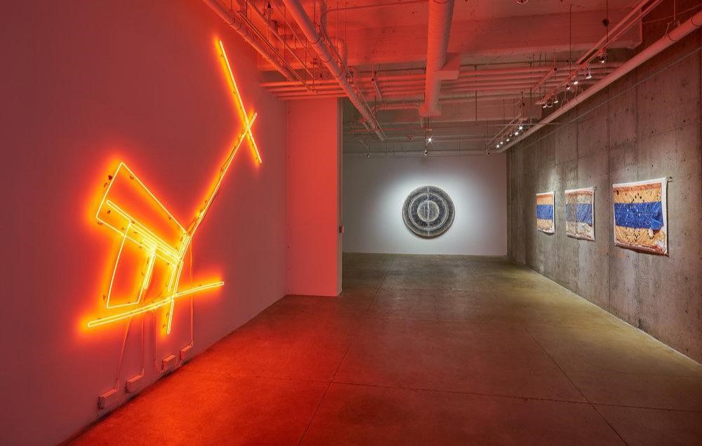 EXHIBITION DOCUMENTATION OF OMAR MISMAR THE PATH OF LOVE #03 , 2013-2014, NEON, 126 X 126 INCHES (LEFT), TARANEH HEMAMI RECOUNTING , 2011, PIGMENT AND INKJET PRINTS ON PAPER AND VELLUM, WAX, WOOD PANELS, 66 INCHES DIAMETER, 7 INCHES DEEP (CENTER) AND GUILLERMO GALINDO SIGUIENDO LOS PASOS DEL NIÑO PERDIDO, 20 MILES EAST OF LAREDO , AND CARTOGRAFÍA DEL ESPIRITU/CARTOGRAPHY OF THE SPIRIT, ALL 2017, ACRYLIC ON BEACON FLAGS USED BY HUMANITARIAN AID GROUP WATER STATIONS (RIGHT). IMAGE COURTESY OF THE ARTISTS AND GALLERY WENDI NORRIS, SAN FRANCISCO.