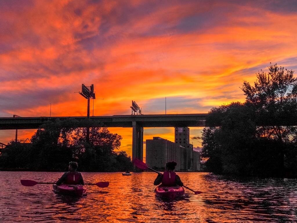  Two kayakers paddle into the sunset on the Menomonee River, with pink, yellow and purple skies ahead of them 