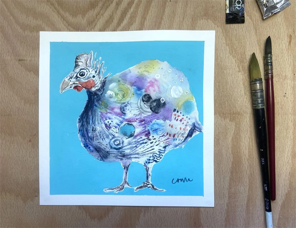A Little Bit Dotty, original watercolour painting of a quirky guinea fowl against a bright turquoise background, photographed on a wooden desk beside paintbrushes.