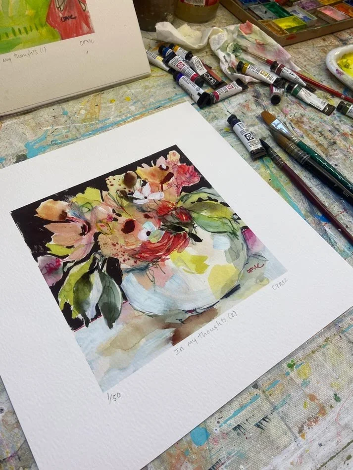 Abundance floral Giclée print on the artist’s studio table, showing expressive pink, peach and yellow flowers in a white vase.