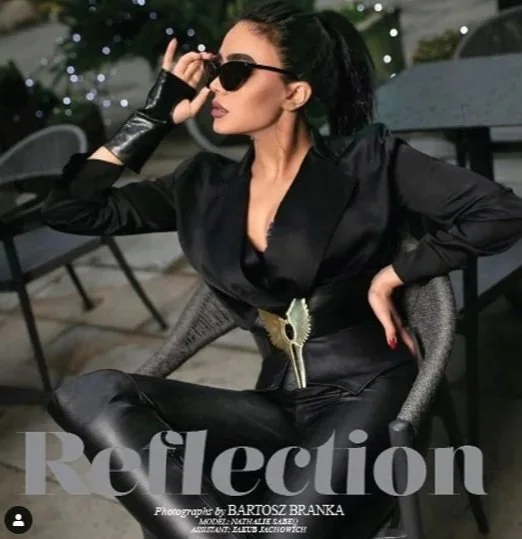 A woman with dark hair tied back, wearing sunglasses, a black satin jacket, and black leather pants, sitting outdoors on a patio with string lights in the background, posing with her hand adjusting her sunglasses.