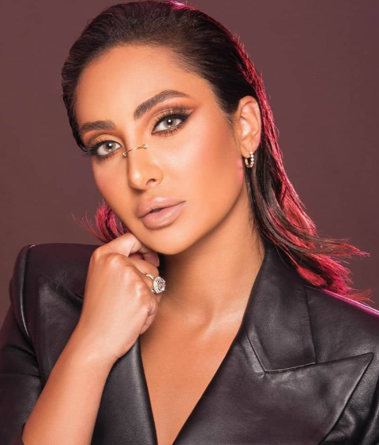 A woman with dark hair styled back, wearing makeup with bold eye makeup and nude lipstick, dressed in a black leather jacket, and wearing earrings, rings, and a delicate nose ring, poses against a plain background.